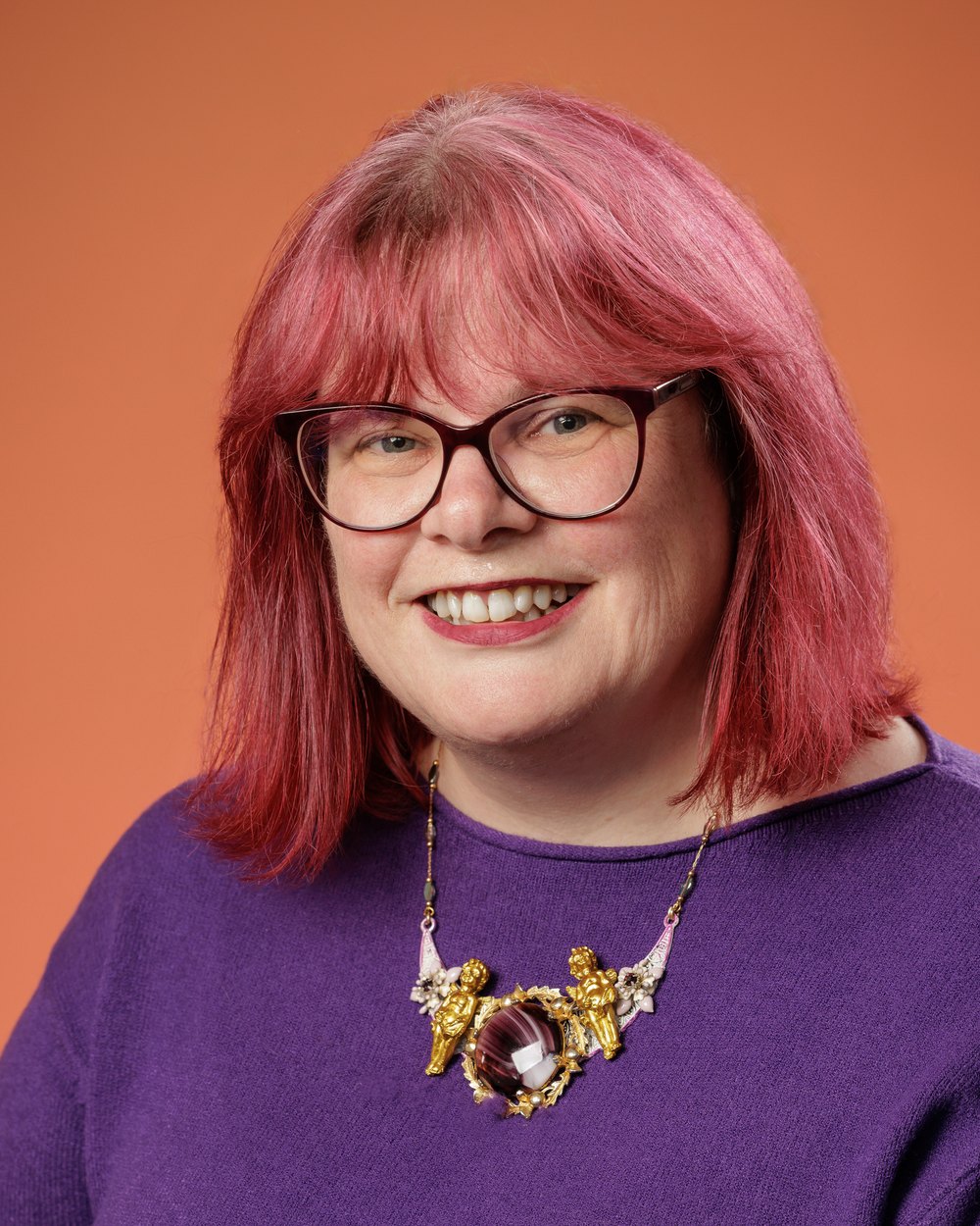 Gill Christie, smiling, with purple hair and glasses, wearing a purple jumper and an ornate gold necklace